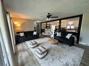 A living room with two sofas, a hanging chair, ottomans, a ceiling fan, and a kitchen in the background.