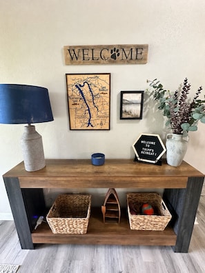 A wooden console table with a map, a welcome sign, a framed picture, a vase with plants, and baskets.