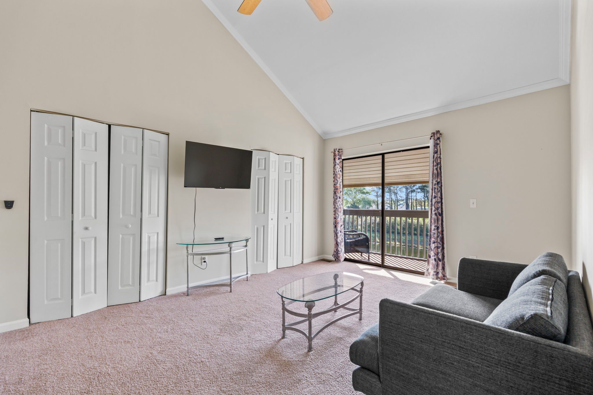 A living room with a sofa, a glass coffee table, a TV mounted on the wall, and a sliding glass door leading to a balcony.