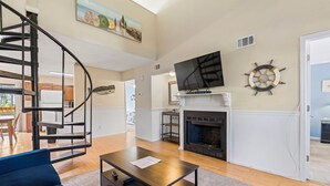 A living room with a fireplace, a TV mounted above it, a staircase, and a dining area in the background. - Surfside Beach Retreat - Pool Access - 4BR - 1 Mile to Beach (Myrtle Beach)
