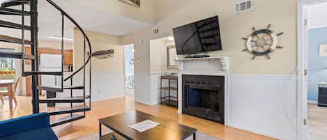 A living room with a fireplace, a TV mounted above it, a staircase, and a dining area in the background.