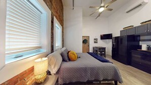 A bedroom with a bed, a bedside lamp, a ceiling fan, and a kitchen area with appliances.
