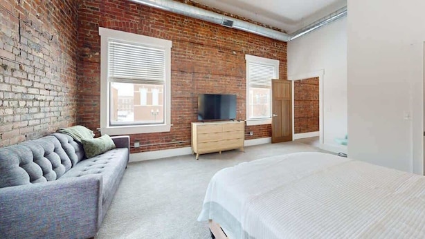 A bedroom with a brick wall, a sofa, a bed, a dresser, and a television.