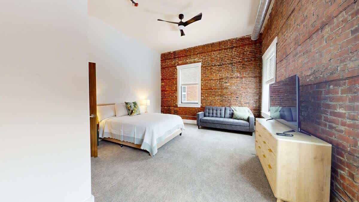 Une chambre avec un mur en briques, un lit, un canapé gris, une commode en bois et un téléviseur.