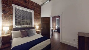 Un dormitorio con pared de ladrillo, un ventanal con persianas, una cama con almohadas y una mesita de noche con lámpara.