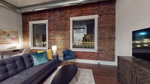 Una sala de estar con pared de ladrillo, dos ventanas amplias, un sofá, una mesa de centro, una mesita auxiliar, una silla y un televisor sobre un mueble de madera.