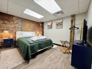 A bedroom with a brick wall, a bed with green bedding, a TV, and artwork on the wall.