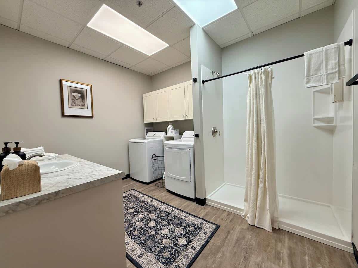 A bathroom with a shower, a toilet, a sink, and a mirror. There is a towel rack above the sink and a framed picture on the wall.