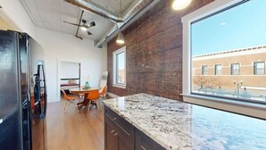 Una cocina con encimera de granito, comedor con mesa y sillas, y un amplio ventanal con vista a un edificio de ladrillo.