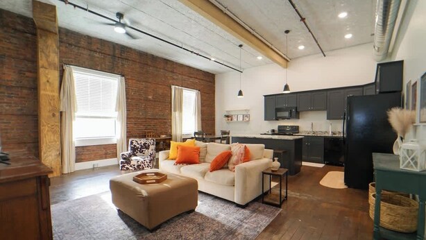 Ein modernes Wohnzimmer mit Sofa, Hocker und einem kleinen Tisch. Es gibt eine Küchenzeile mit Schränken und einem Kühlschrank.
