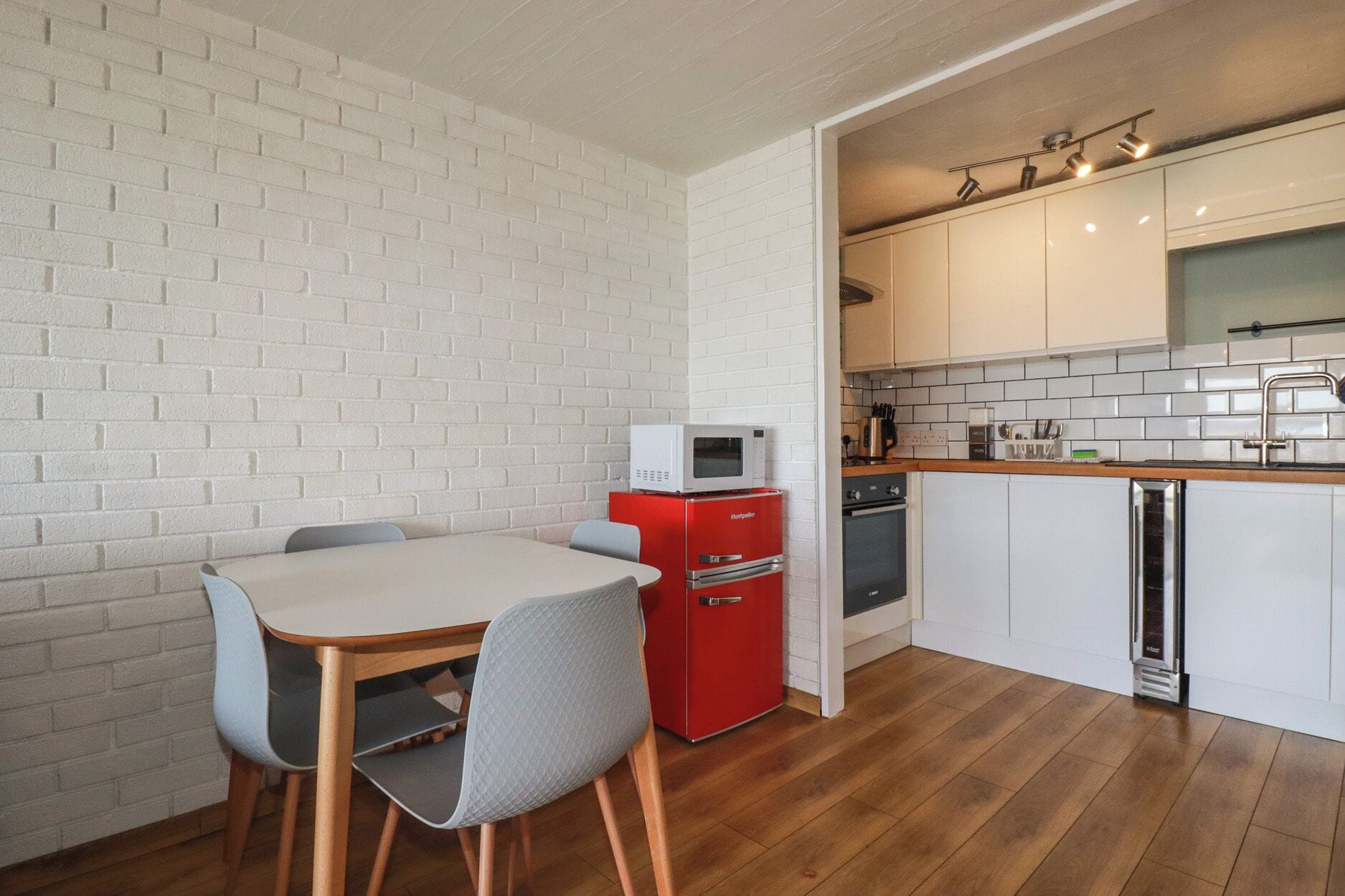 Una cocina compacta con paredes de ladrillo blanco, un electrodoméstico rojo y una zona de comedor con mesa y sillas.