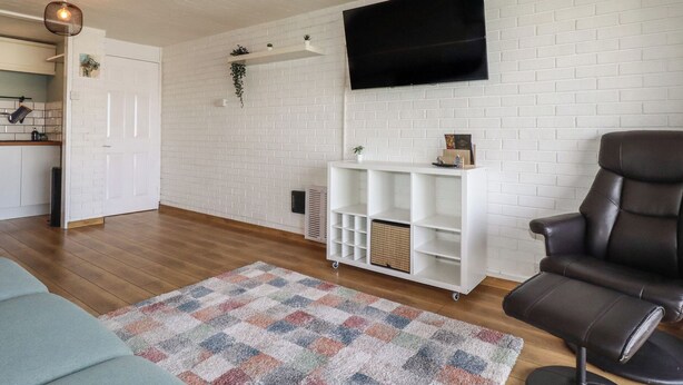 Una sala de estar con televisor, sofá, sillón, alfombra estampada y pared de ladrillo blanco.