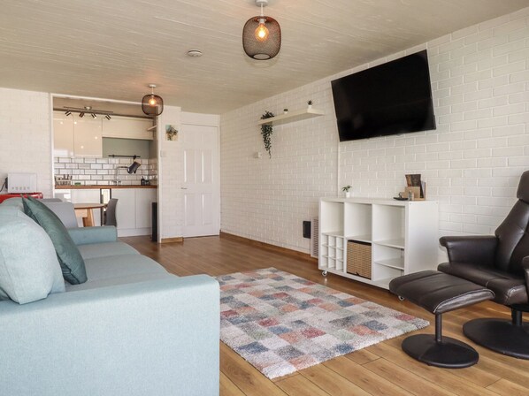 A modern living room with a sofa, a leather chair, a TV, and a kitchen in the background. - Fistral Bay Apt (Newquay)