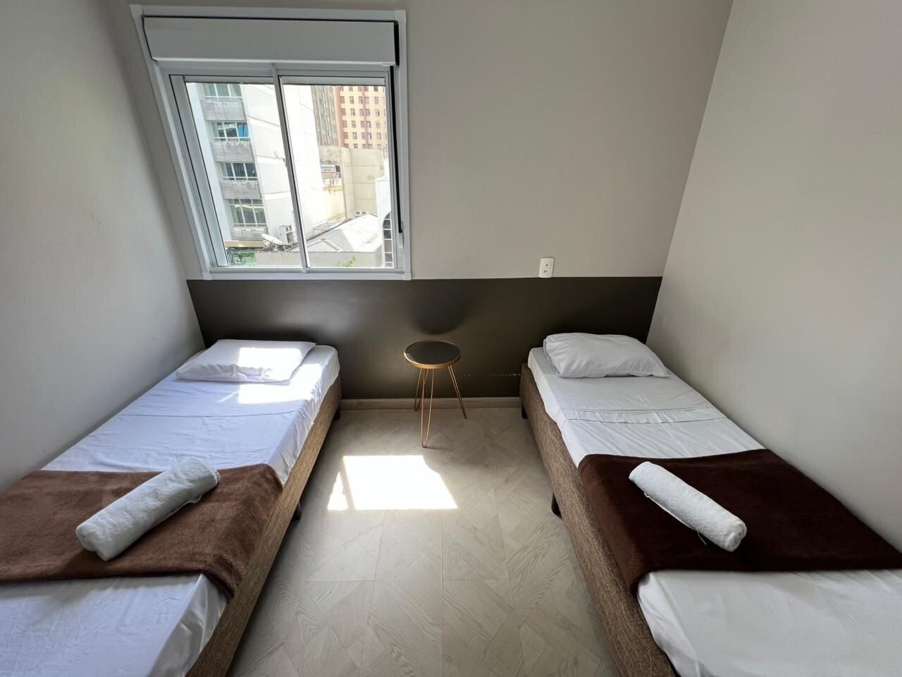 A small, well-lit room with two single beds, a small round table, and a window providing a view of buildings outside.