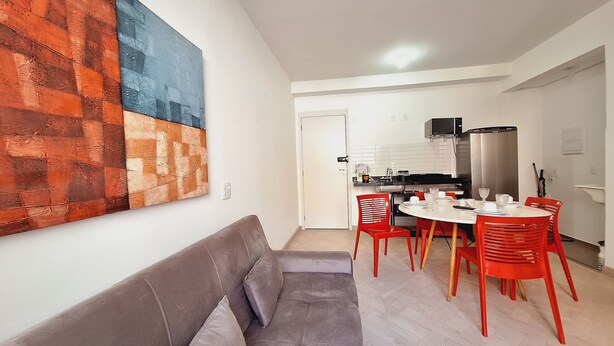 A modern living room with a grey sofa, red chairs, and a dining table. A kitchen is visible in the background with white cabinets and a stainless steel refrigerator. - LK Centro 2 (Belo Horizonte)
