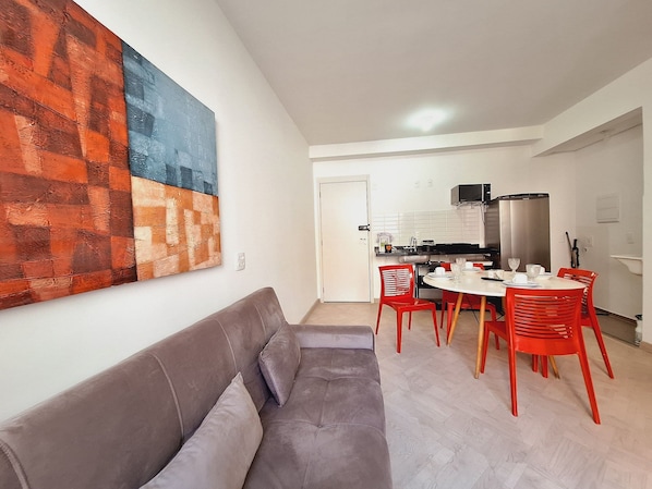 A modern living room with a grey sofa, red chairs, and a dining table. A kitchen is visible in the background with white cabinets and a stainless steel refrigerator.