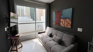 A modern living room with a grey sofa, a small wooden side table, a TV, and a balcony with a view of buildings. - LK Centro 2 (Belo Horizonte)