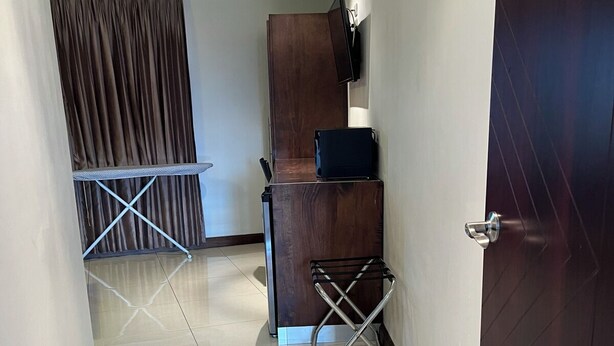 A hallway with a wooden door, a dark wooden cabinet, a small black TV on the wall, and a metal folding chair. - Paria Suites Hotel and Conference Center (La Romain)