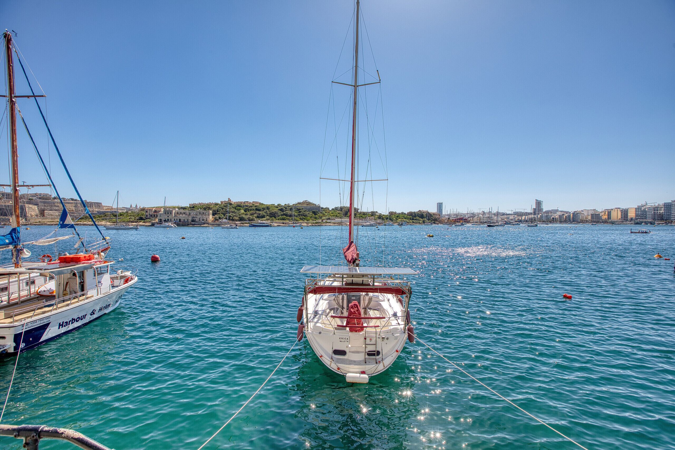 Un port amb un veler amarrat, altres embarcacions i un paisatge urbà al fons.