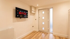 Huoneessa on seinälle asennettu taulutelevisio, josta näkyy Netflixin sisältöä, ovi, jossa on himmeät lasipaneelit, ja puulattia.