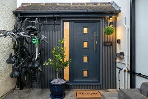Una puerta oscura con felpudo, una planta en maceta y bicicletas estacionadas afuera.