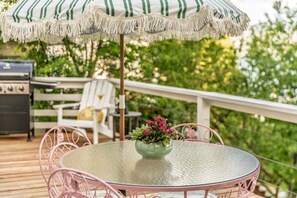 Et rundt glasbord med en grøn vase med lyserøde blomster, en lyserød metalstol og en stribet parasol på en træterrasse.