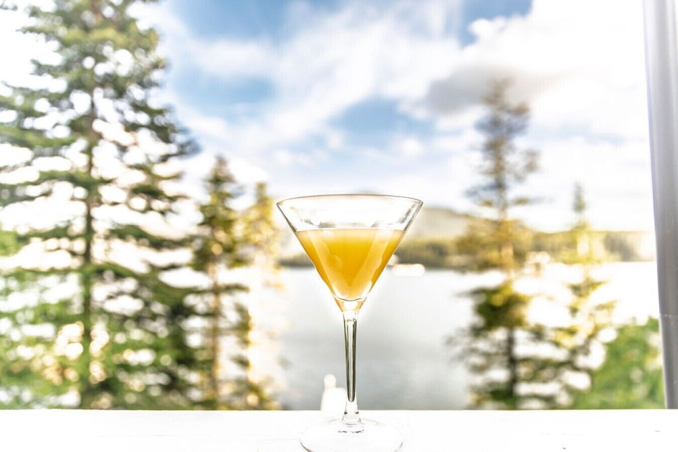 A martini glass with a clear liquid on a table with a view of trees and a cloudy sky.