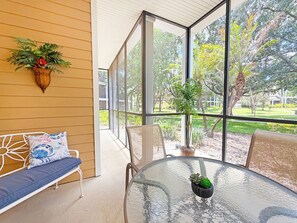 Ein Zimmer mit Glastisch, einer Bank mit Kissen, einer Topfpflanze und Blick auf einen Garten durch große Fenster.