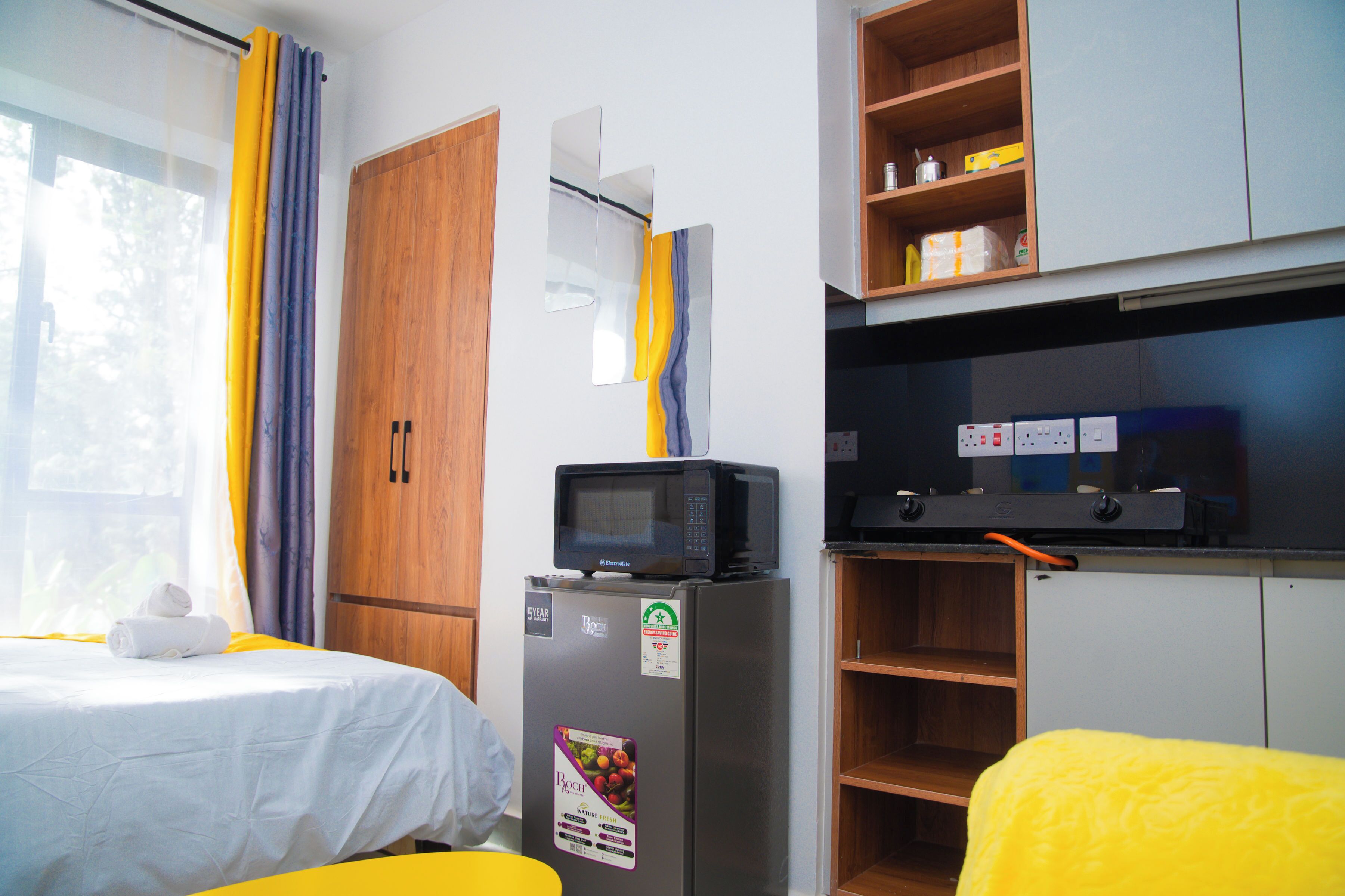 A hotel room with a bed, a refrigerator, a microwave, a wooden wardrobe, and a kitchenette area.