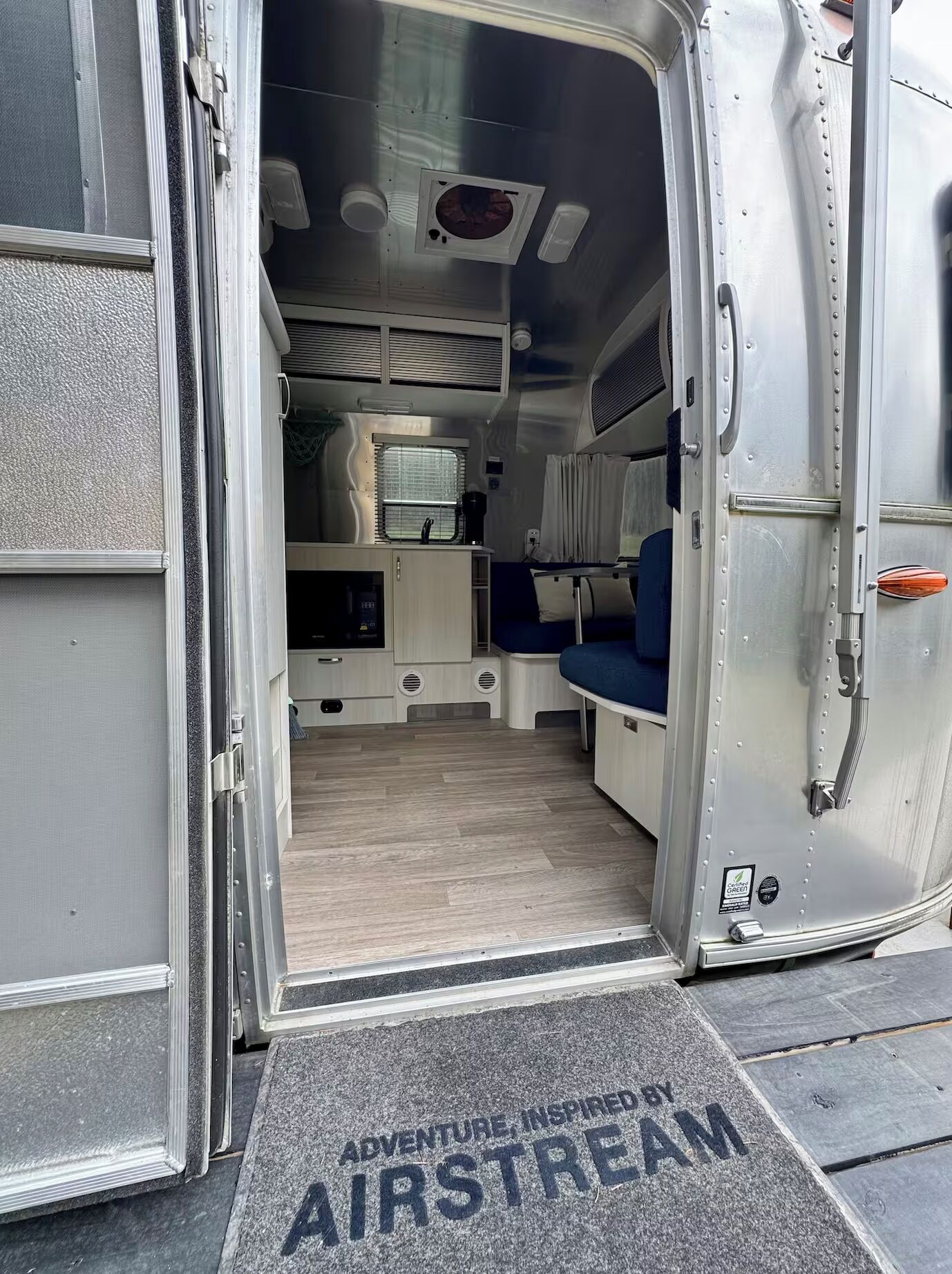 An open van with a kitchen area, a sleeping berth, and a door mat with 'Adventure Inspired by Airstream' text.