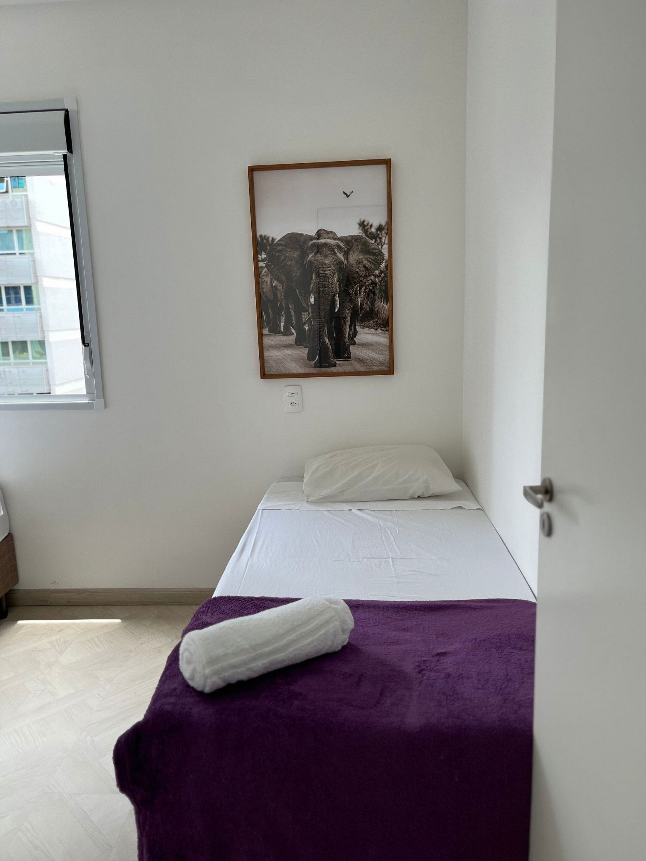 A bedroom with a bed, a purple blanket, a white pillow, and a framed picture on the wall.