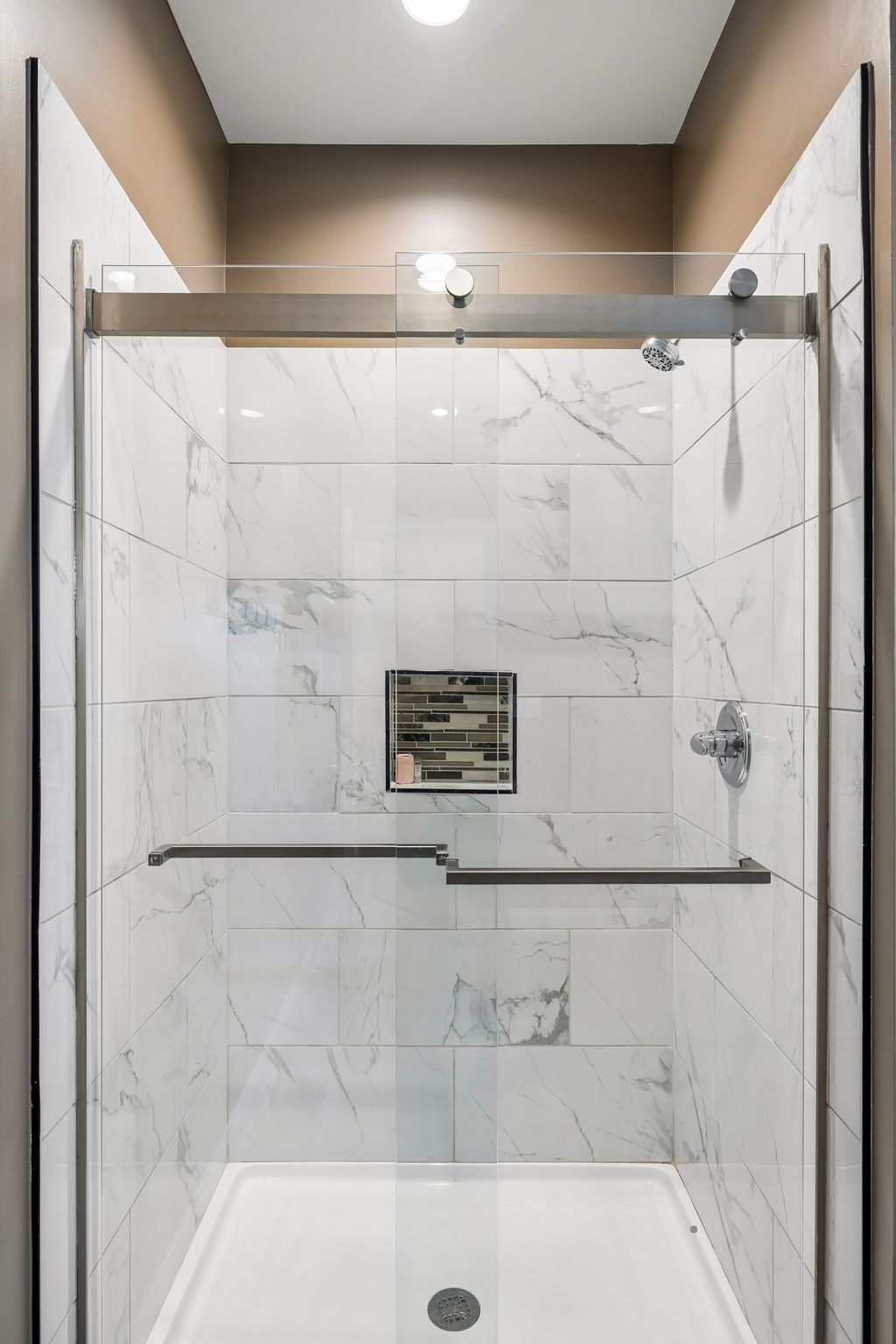 Une douche à l’italienne avec une porte en verre, des carreaux de marbre, une barre d’appui et un luminaire au plafond.