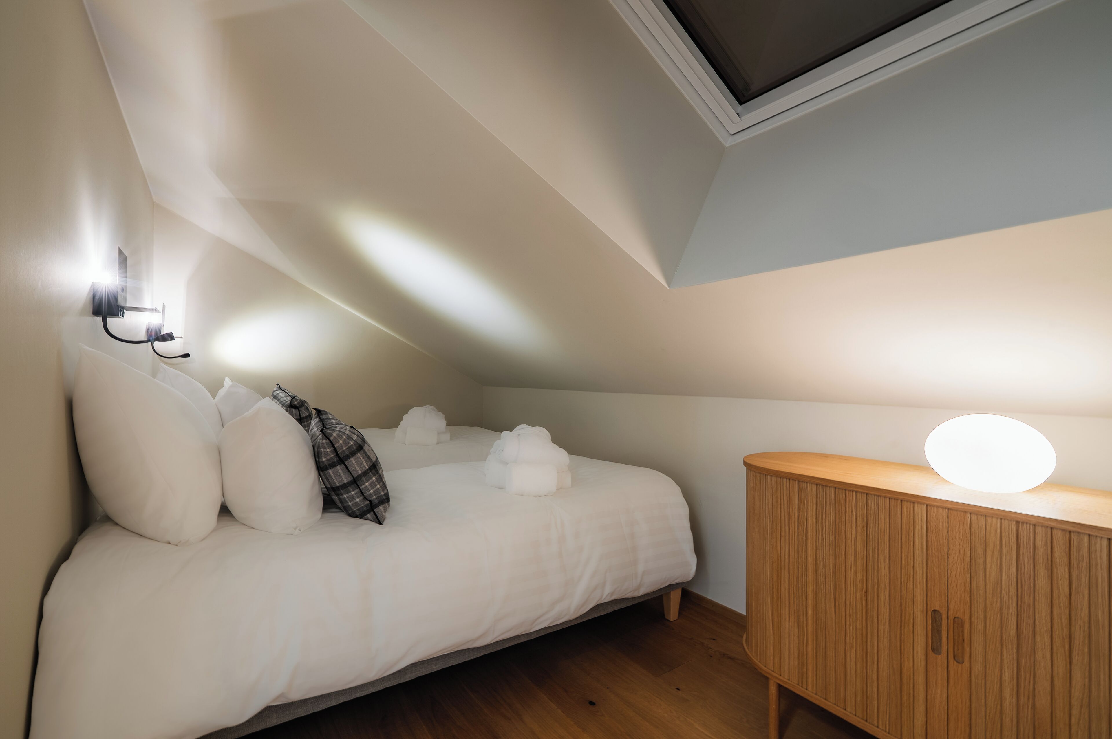 A bedroom with a bed, pillows, a bedside table with a lamp, and a skylight.