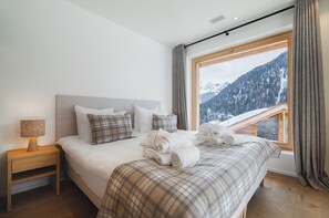 A bedroom with a large bed, a bedside table, a lamp, and a window with a mountain view. - Guerneres Resort & Spa (Grimentz)