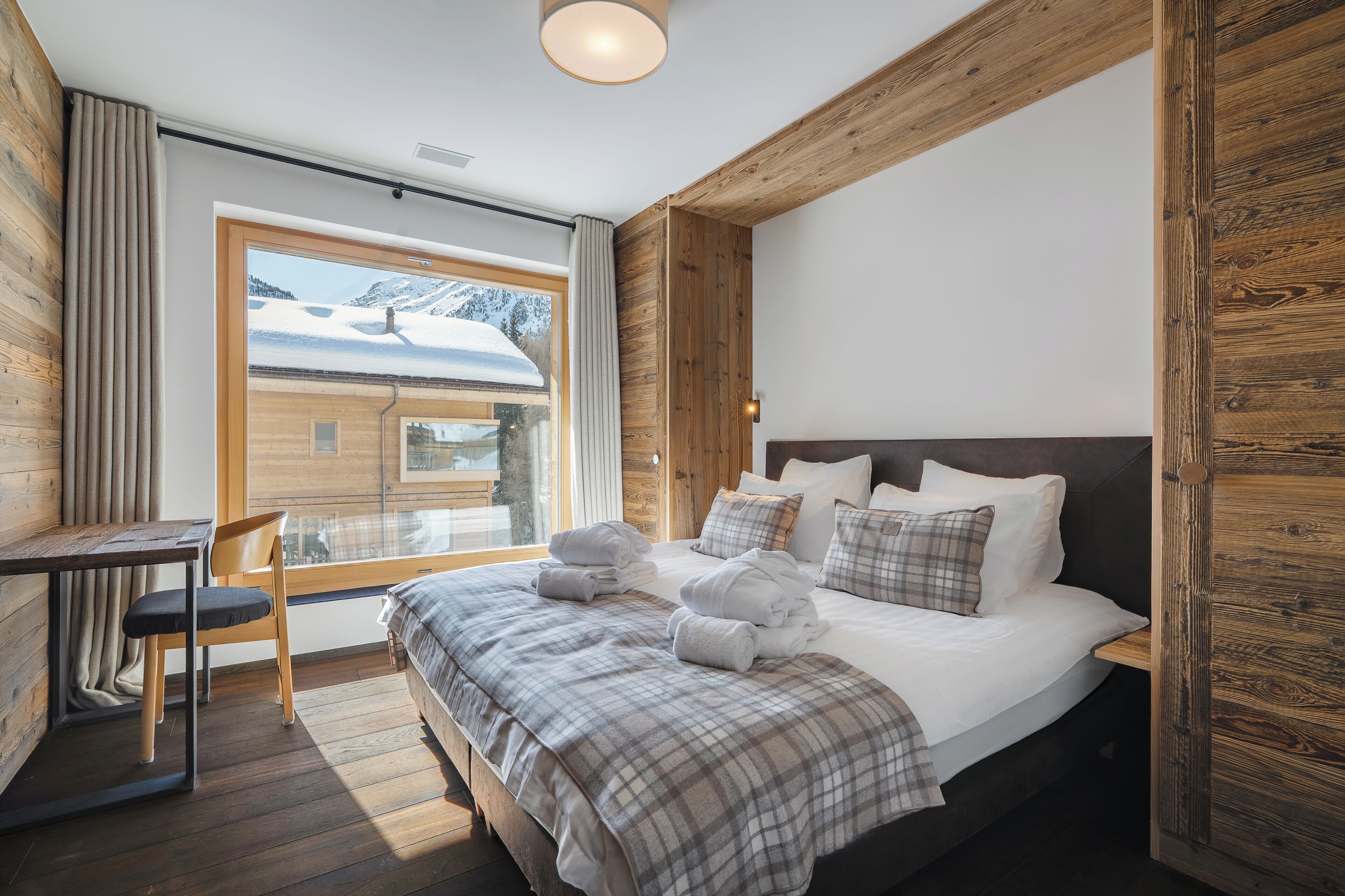 A bedroom with a large bed, a wooden headboard, a window with a view of snow-covered mountains, and a small table with a chair.
