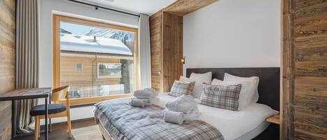 A bedroom with a large bed, a wooden headboard, a window with a view of snow-covered mountains, and a small table with a chair.