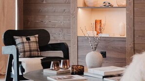 A modern living room with a wooden wall, a black chair with a plaid cushion, a round coffee table with books and decorative items, and a shelf with various objects. - Guerneres Resort & Spa (Grimentz)