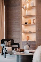 Uma sala de estar moderna com uma parede de madeira, uma cadeira preta com almofada xadrez, uma mesa de centro redonda com livros e objetos decorativos, e uma estante com diversos objetos.