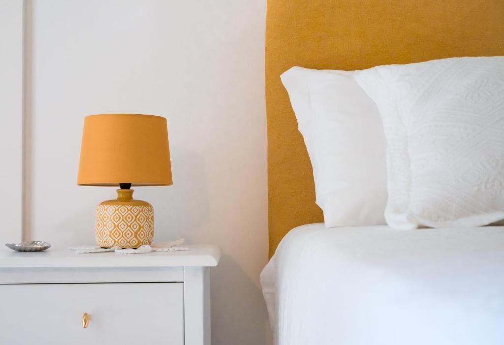 Una cama bien hecha con ropa blanca, una mesita de noche blanca con una lámpara estampada y una pared lisa.