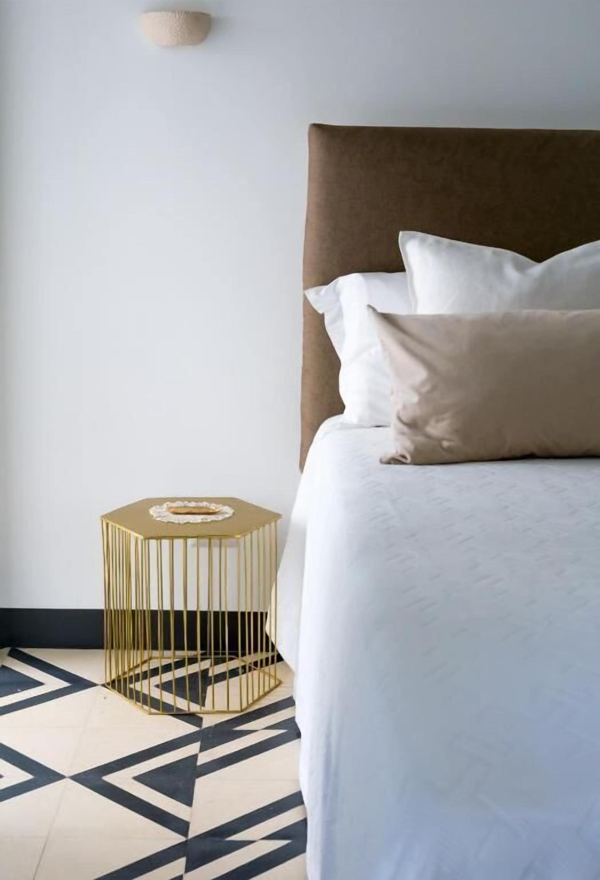 A bed with a brown headboard and white bedding, a gold and black patterned floor, and a gold side table with a white object on it.