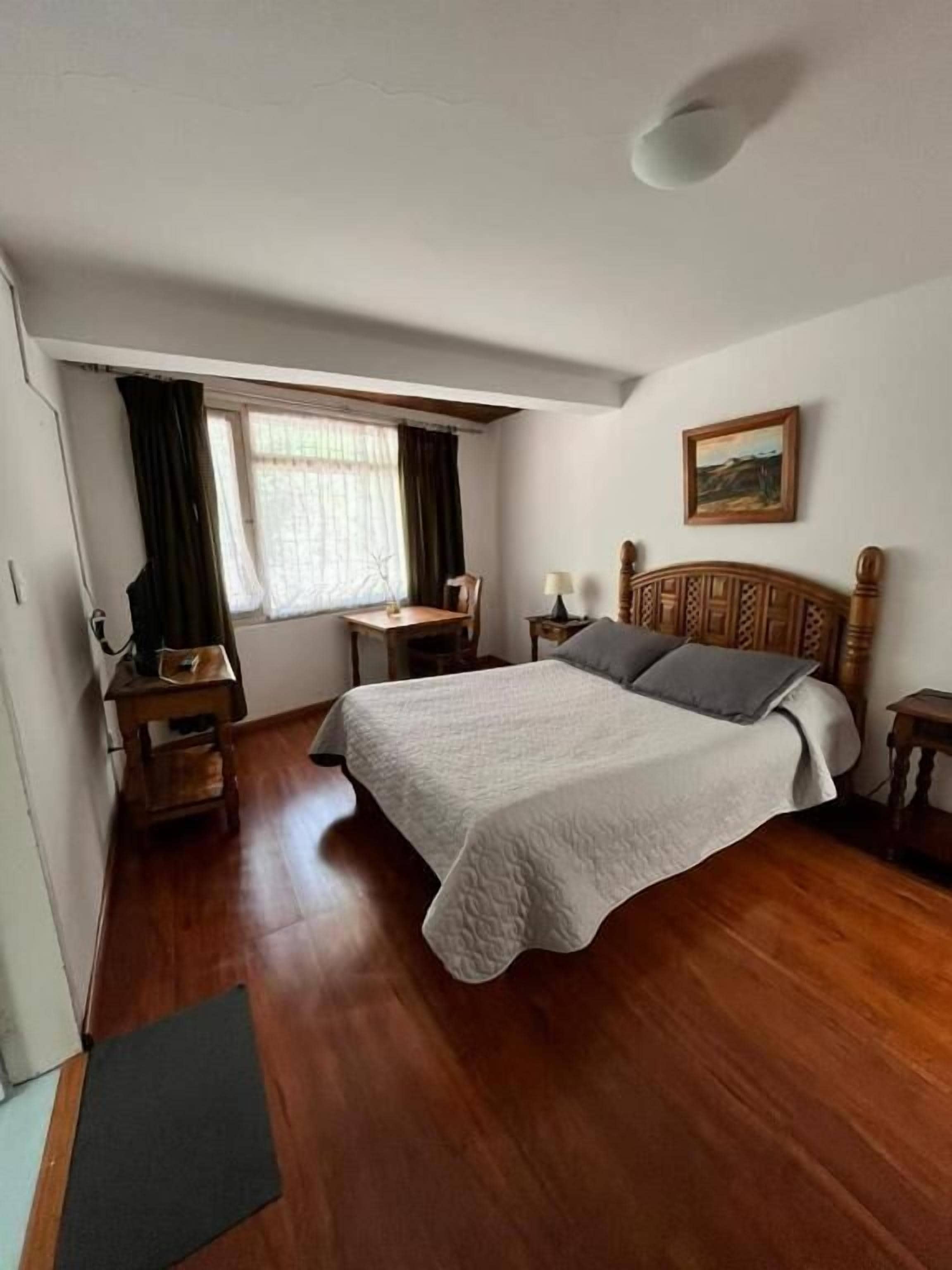 Quarto com cama de madeira, uma escrivaninha, cadeira, mesa de cabeceira, janela com cortinas e um quadro na parede.