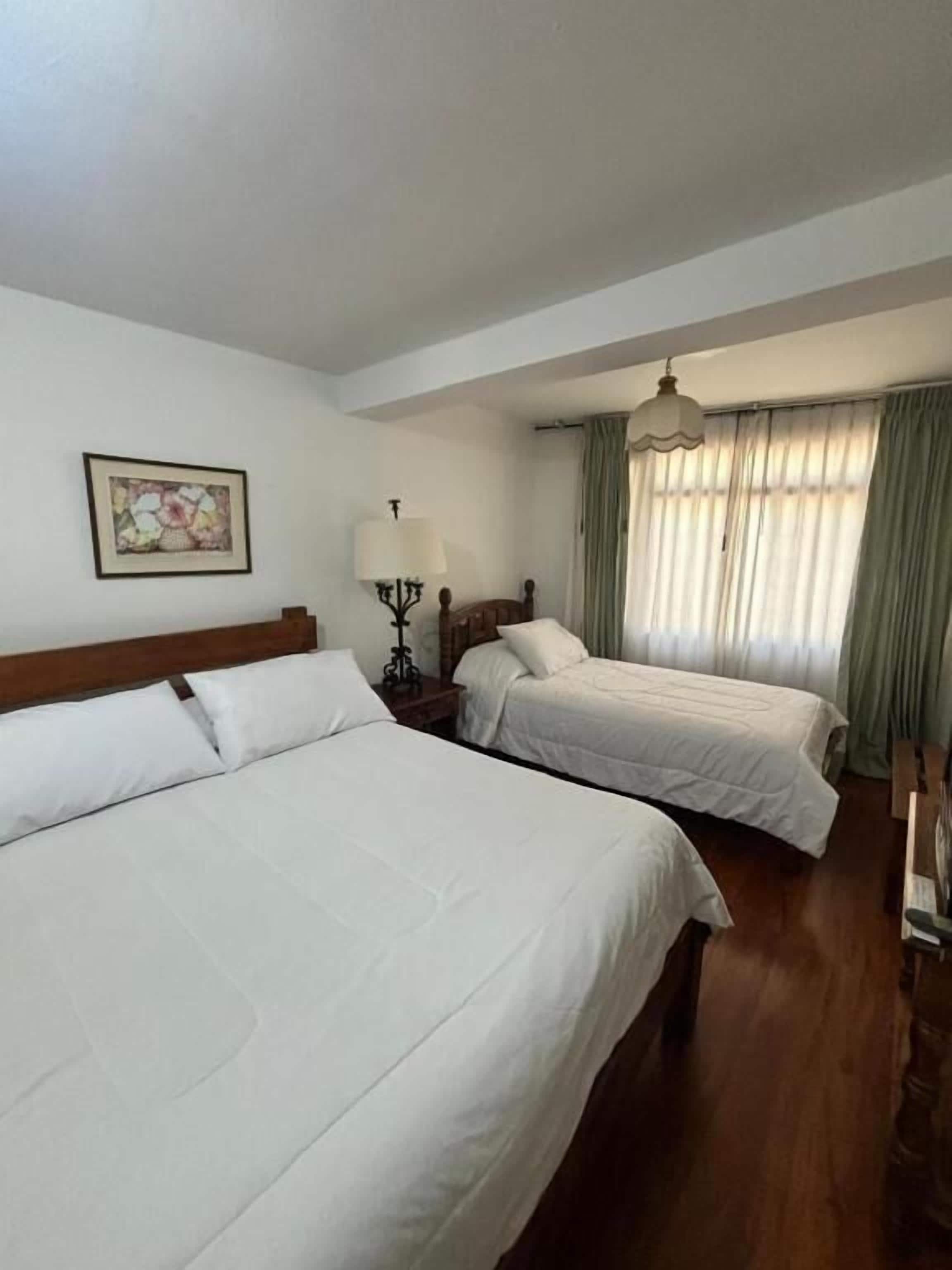 A bedroom with two beds, a nightstand, a lamp, a painting on the wall, and a window with curtains.