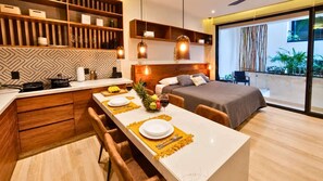 Ein modernes Hotelzimmer mit Küchenbereich, einem Esstisch für zwei Personen, einem Bett und einem Balkon mit Pflanzen.