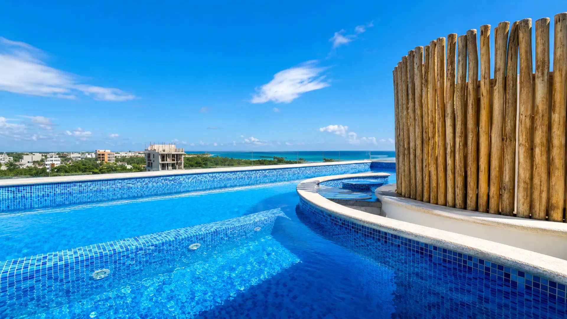 Ein klarer, blauer Pool mit Holzzaun, Blick aufs Meer und ein Gebäude im Hintergrund.