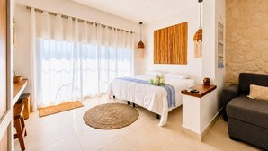 A bedroom with a bed, a bedside table, a sofa, a large window with curtains, a wall-mounted light fixture, and a woven rug. - Bright Balcony Studio Beach Vibe · Blanko Living (Playa del Carmen)