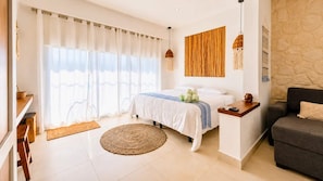 A bedroom with a bed, a bedside table, a sofa, a large window with curtains, a wall-mounted light fixture, and a woven rug. - Bright Balcony Studio Beach Vibe · Blanko Living (Playa del Carmen)