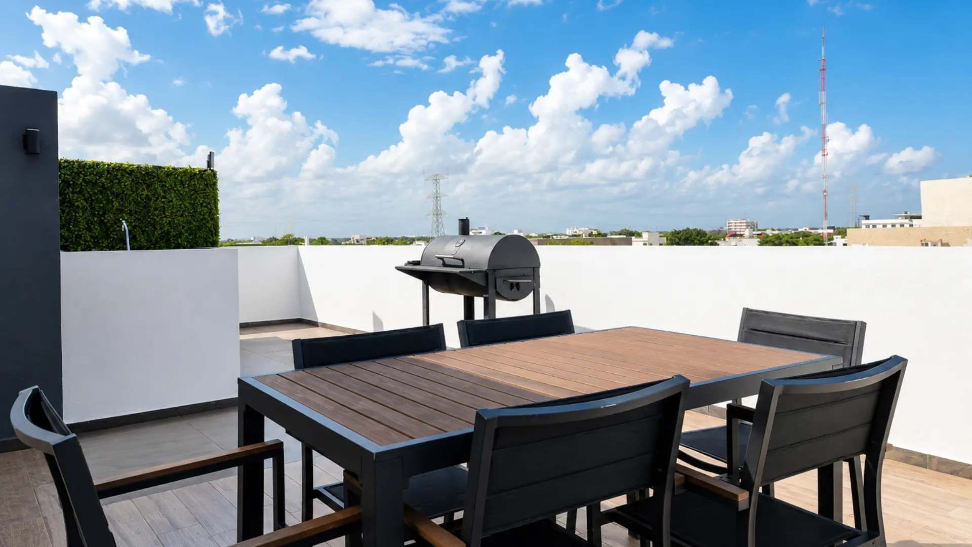 A rooftop patio with a wooden table and chairs, a grill, and a clear view of the city skyline.