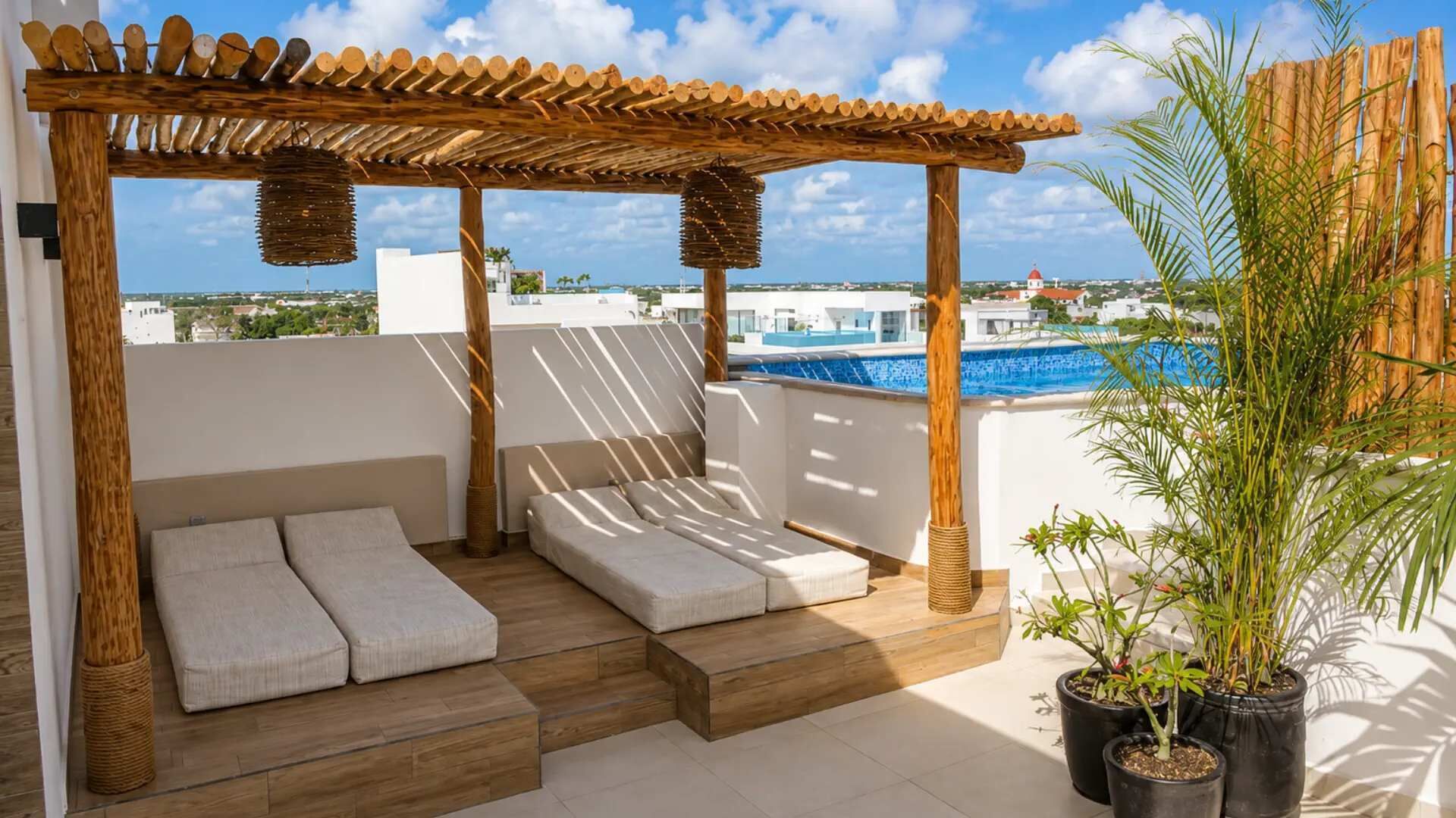 Une terrasse sur le toit agrémentée d’une pergola en bois, de deux coins salon avec des coussins, d’une piscine et de plantes en pot.