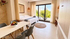 Ein Hotelzimmer mit Bett, Esstisch mit Stühlen, einem kleinen Balkon mit Sitzgelegenheit und Blick auf Grünflächen und Gebäude.