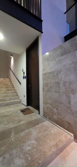 Eine Treppe führt zu einer Tür mit Fußmatte, einer Handläufung und einer Wand mit strukturierter Oberfläche.